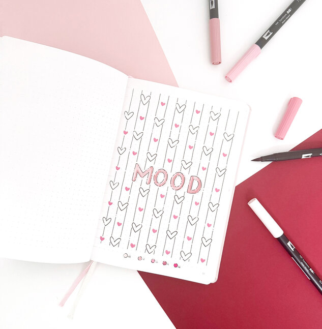 Open bullet journal with a creatively designed mood tracker. The page contains the word "MOOD" in capital letters, surrounded by vertical lines with drawn pink hearts, as well as outlines of hearts that can be colored according to your mood. At the bottom of the page is a mood scale with different shades of pink.